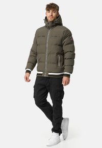 Dark green puffer jacket with a high collar, zippered chest pockets, white accents, and elastic cuffs, worn with black cargo pants and white sneakers.