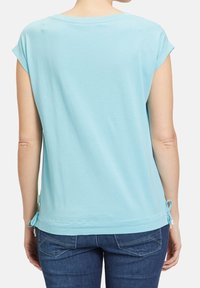 Vista trasera de una persona que lleva una blusa de manga corta azul claro con lazos laterales y jeans azul oscuro frente a un fondo neutro.