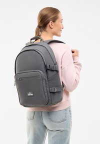 Rucsac gri realizat dintr-un material durabil, având un buzunar frontal cu fermoar, curele ajustabile și un spate căptușit. Design simplu, modern, cu o textură moale.