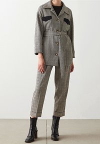 Kostkovaná bunda s černými kapsami, knoflíkovým zapínáním a opaskem. Doplněno o kostkované kalhoty a černé kotníkové boty. Hladká textura.