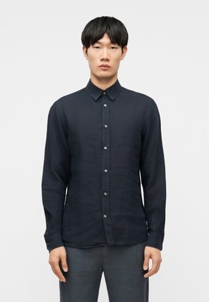 Νεαρός άνδρας με μαύρα μαλλιά, φορώντας σκουρό μπλε πουκάμισο με κουμπιά και γκρι παντελόνι, στέκεται μπροστά σε ένα απλό ανοιχτόχρωμο φόντο.