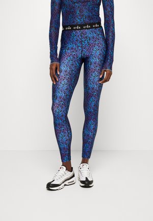 Eine Person steht in lebhaft blauen und lila gemusterten Leggings und einem Langarmshirt, trägt weiße und schwarze Sportschuhe.
