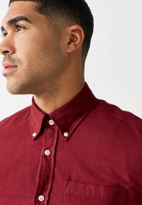 Chemise bordeaux boutonnée en tissu texturé, dotée d'un col pointu, de boutons blancs et d'une poche poitrine sur le côté gauche.