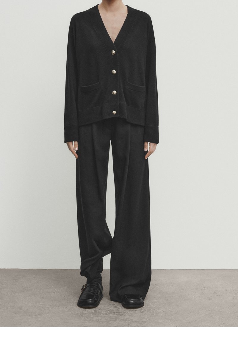 Personne portant un cardigan noir ample boutonné et un pantalon noir à jambes larges avec des chaussures noires, debout contre un mur blanc uni.