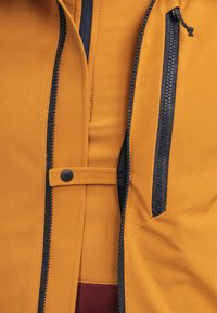 Gelbe Jacke mit schwarzen Reißverschlussverschlüssen, einem Druckknopf und einer glatten Textur, unter der ein weinrotes Futter sichtbar ist.