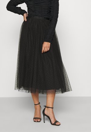 Frau trägt ein schwarzes, langärmliges Oberteil mit Raffung, einen schwarzen, transparenten Midirock mit Sternenmuster und schwarze Sandalen mit hohen Absätzen und Knöchelriemen.