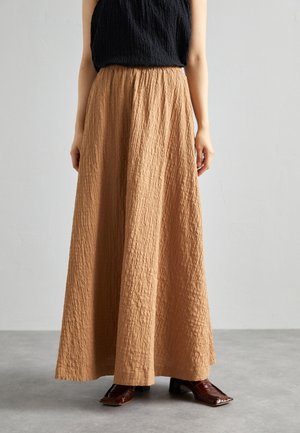 Frau trägt ein schwarzes ärmelloses Oberteil, langen strukturierten braunen Rock und dunkelbraune Schuhe mit quadratischer Spitze, steht auf einem grauen Boden.