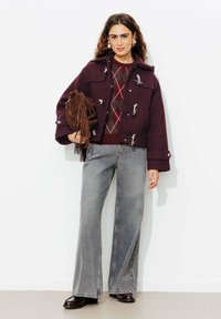 Veste en laine bordeaux avec des fermetures à bascule sur un pull à motif en losanges, associée à un jean gris évasé et un sac à franges marron.
