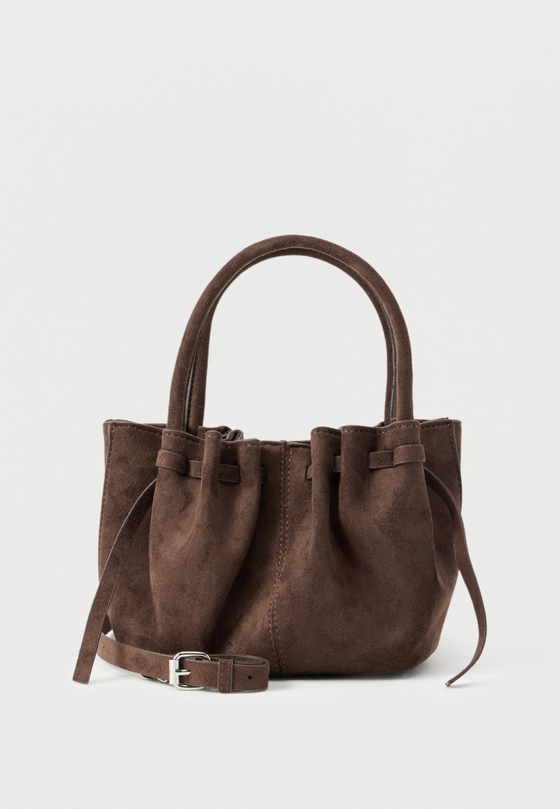 Καφέ τσάντα χειρός από σουέτ με στρογγυλεμένο πάνω μέρος, διπλές λαβές και αποσπώμενο ιμάντα με ασημένια πόρπη, παρουσιάζεται σε απλό φόντο.