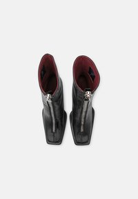 Paire de bottines en cuir noir avec des fermetures éclair à l’avant et une doublure intérieure bordeaux, vues de dessus sur un fond blanc.