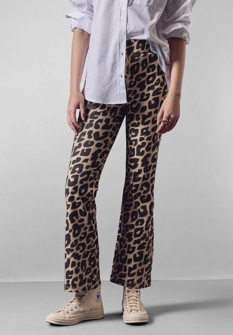 Kostkované kalhoty ve zvířecím vzoru s rozšířenými nohavicemi v béžové a černé barvě, vyrobené z hladkého materiálu. Doplňeny s bílou košilí na knoflíky a béžovými teniskami.