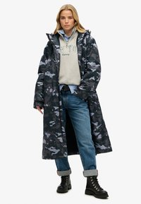 Długi czarny i szary płaszcz w kamuflażu z kapturem, noszony na szarym swetrze i niebieskich dżinsach, w połączeniu z czarnymi butami na sznurówki.