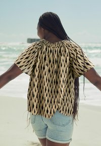 Haut beige avec un motif ikat noir, encolure froncée, et manches courtes, associé à un short en denim clair, sur fond de plage.