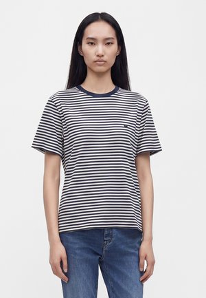 Jeune femme aux longs cheveux foncés, portant un t-shirt à manches courtes rayé bleu marine et blanc et un jean bleu, debout devant un fond blanc uni.