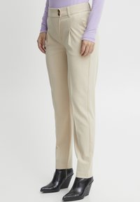 Pantaloni beige su misura con due pieghe frontali, chiusura con un singolo bottone marrone e vestibilità slim, abbinati a stivaletti neri con tacco a cuneo.