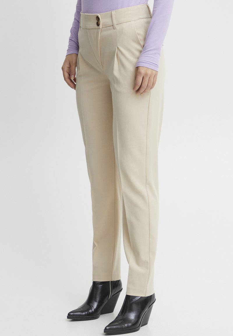 Pantalons tailleur beiges avec deux plis sur le devant, une fermeture à un bouton marron, et une coupe ajustée, associés à des bottines à talons compensés noires.