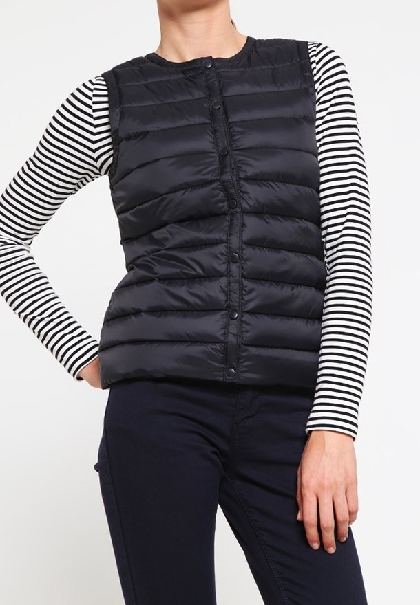 Person som bär en svart pufferväst över en svart-vit randig långärmad skjorta och mörka byxor, stående med ena handen på höften.