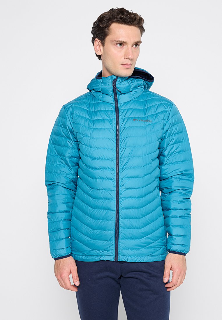 Mężczyzna w tealowej, pikowanej kurtce Columbia z kapturem, stojący na tle jasnego, jednolitego tła, patrzący w bok.