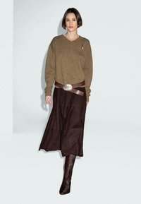 Una donna con i capelli corti e scuri indossa un maglione marrone a scollo a V, una gonna larga scura, una cintura larga marrone e stivali alti marroni, mentre sta in piedi contro un muro bianco.