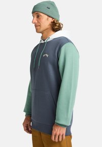 Hoodie w kolorze granatowym i jasnolimonkowym, z kieszenią z przodu, kapturem ze sznurkiem i haftowanym logo na piersi. Tekstura materiału.