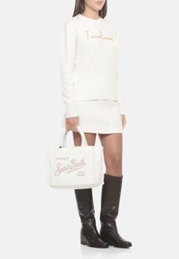 Pull blanc à manches longues avec « Fawless » en fil doré ; mini-jupe texturée blanche ; sac fourre-tout blanc avec « Dreaming of Saint Barth » en rose pâle ; bottes marron montant jusqu'aux genoux.