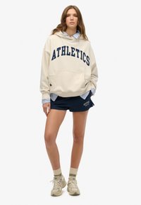 Cream hoodie with "ATHLETICS" in navy, front pocket; paired with navy shorts featuring logo. Model wears beige sneakers with green accent socks.