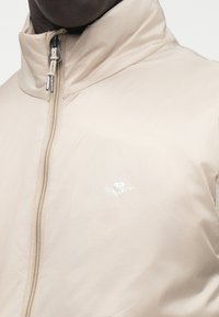 Bež lagana jakna s visokim ovratnikom, s prednjim patentnim zatvaračem i malim bijelim logotipom na prsima. Glatka tekstura.