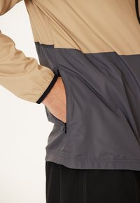 Leichte beige und graue Jacke mit seitlicher Zipper-Tasche und elastischen Bündchen, gefertigt aus einem glatten, leicht strukturierten Stoff.