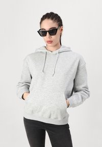 Gray hooded sweatshirt made of soft fabric, featuring a front pouch pocket and adjustable drawstrings. Worn with black pants and sunglasses.