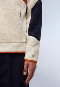 Cream and navy fleece jacket with textured fabric, orange accents on cuffs and hem, featuring a side pocket and logo detail on sleeve.