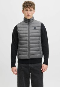 Pelēks pūkains vāciņš ar augstu apkakli, horizontālu quilted rakstu, priekšējo rāvējslēdzi un apaļu logotipu, uzvilkts virs melnas džempera.