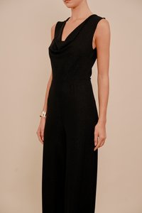 Černý overal bez rukávů s kapucí, přiléhavým pasem a širokými nohavicemi. Vyrobený z lesklého materiálu, zdobený elastickým detailem v pase.