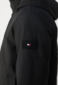 Svart jacka med ett texturerat tyg, en huva och en framträdande svart patch med en röd, vit och blå logotyp på vänster ärm.