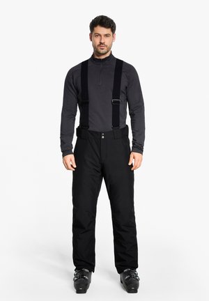 Man wearing black ski pants with suspenders, a dark gray long-sleeve zip-up top, and black ski boots, standing against a white background.