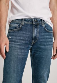 Jeans de mezclilla azul con un corte clásico, ligero desgaste, cinco bolsillos y acentos de costura amarilla. Textura suave.