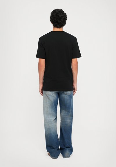 Uomo con capelli ricci che indossa una maglietta nera semplice e jeans blu scoloriti, in piedi di spalle su uno sfondo bianco.
