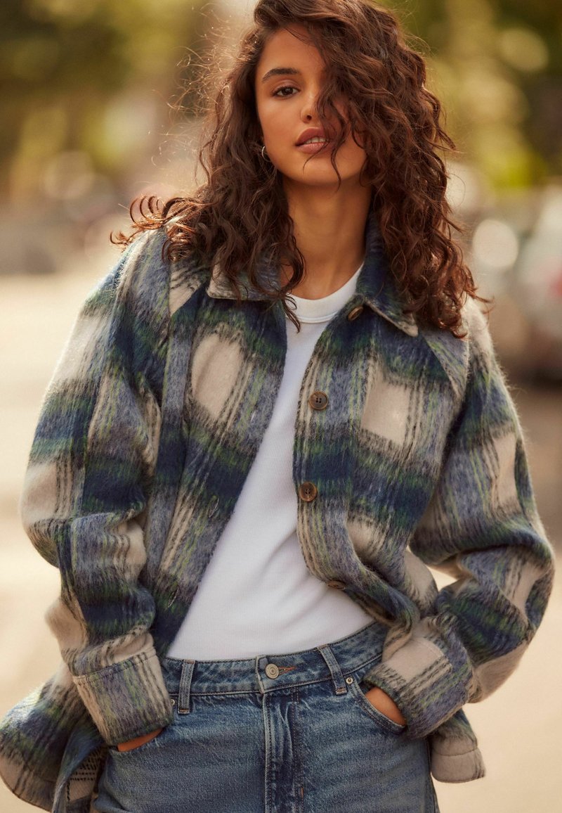 Camicia giacca in misto lana con motivo a quadretti blu, verde e crema; presenta quattro bottoni, colletto e una vestibilità comoda. Abbinata a jeans a vita alta.