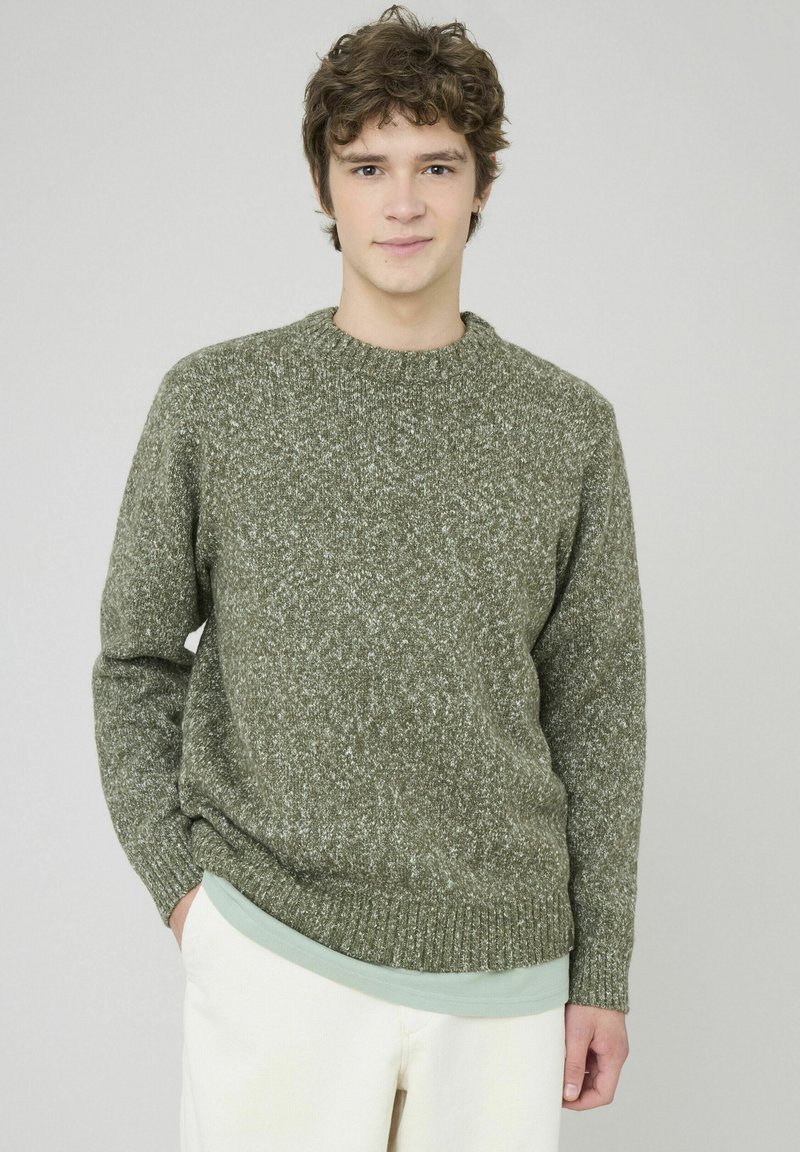 Pull en maille vert olive avec un col rond classique, un tissu texturé et des bords côtelés aux poignets et à l'ourlet, porté par-dessus un sous-vêtement léger de couleur menthe.