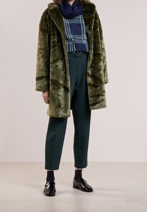 Olivgrön fuskpälsjacka med uppvikta kragar, kombinerad med en blå rutad tröja, gröna byxor och svarta blanka loafers. Slät textur.