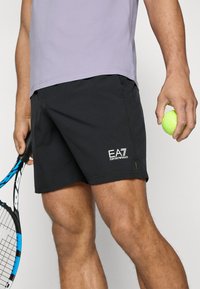 Calções atléticos pretos com um logo na perna esquerda, combinados com uma camisa lilás clara. Uma bola de ténis e uma raquete são seguradas nas mãos.