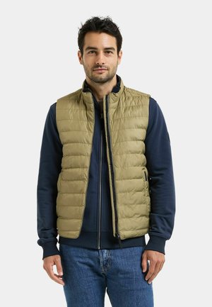 Man staat recht vooruit, draagt een zandkleurig gewatteerd vest over een marineblauwe sweatshirt met rits en blauwe jeans, tegen een effen lichte achtergrond.