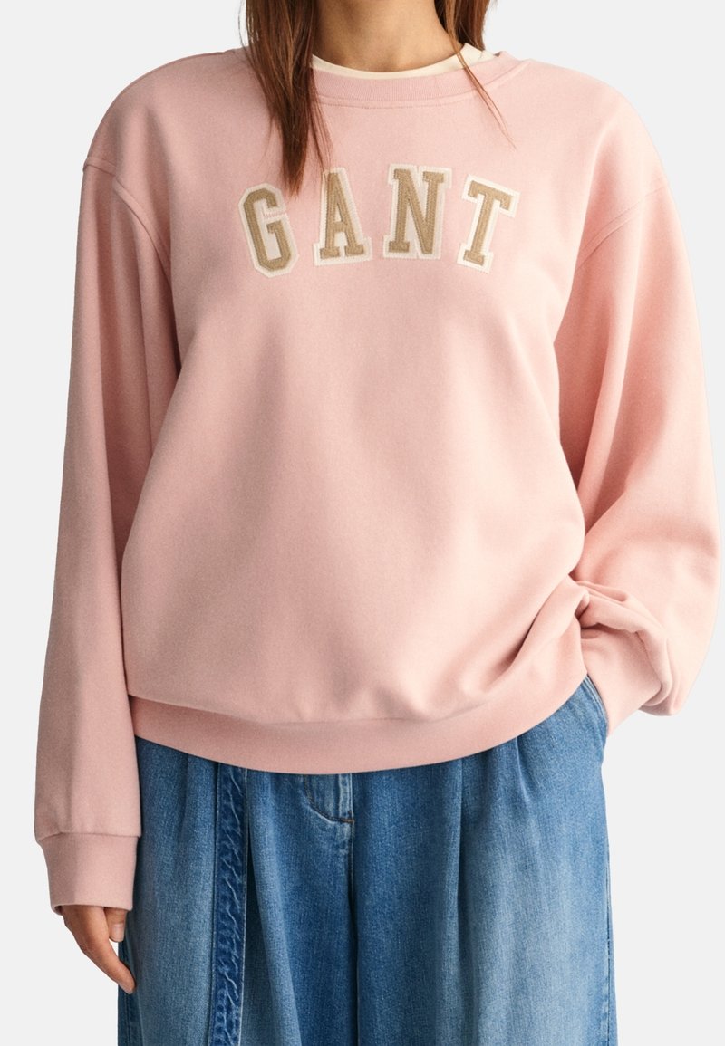 Sweat-shirt rose en tissu doux avec une coupe ample. Porte le mot "GANT" brodé en lettres beige sur la poitrine. Manches longues, poignets côtelés.