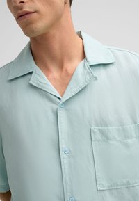 Kurzarmhemd aus leichtblauem Stoff mit einem Schalkragen, einer Brusttasche und vorderer Knopfleiste mit passenden Knöpfen.