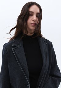 Tmavě modrý denimový blejzr se strukturovanými klopami, zdobený malými cvoky. Nositelný přes dobře padnoucí černý rolák. Hladká textura a přizpůsobený střih.