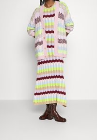 Neuletta, jossa on monivärisiä raitoja pastellisävyissä, kukkakuvioinen vaaleanpunainen resori ja kultaiset napit, yhdistettynä yhteensopivaan raidalliseen mekkoon.