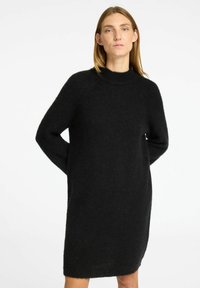 Schwarzes gestricktes Kleid mit hohem Ausschnitt, langen Ärmeln und lockerem Schnitt. Die Textur wirkt weich und kuschelig, was die Wärme erhöht.