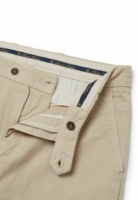 Pantaloni beige in cotone con zip e chiusura a bottone, caratterizzati da una superficie liscia e un'etichetta in vita blu con lettering dorato.