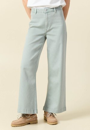 Persona con jeans de pierna ancha azul claro, camiseta blanca y zapatos náuticos beige con cordones, de pie frente a un fondo beige liso.