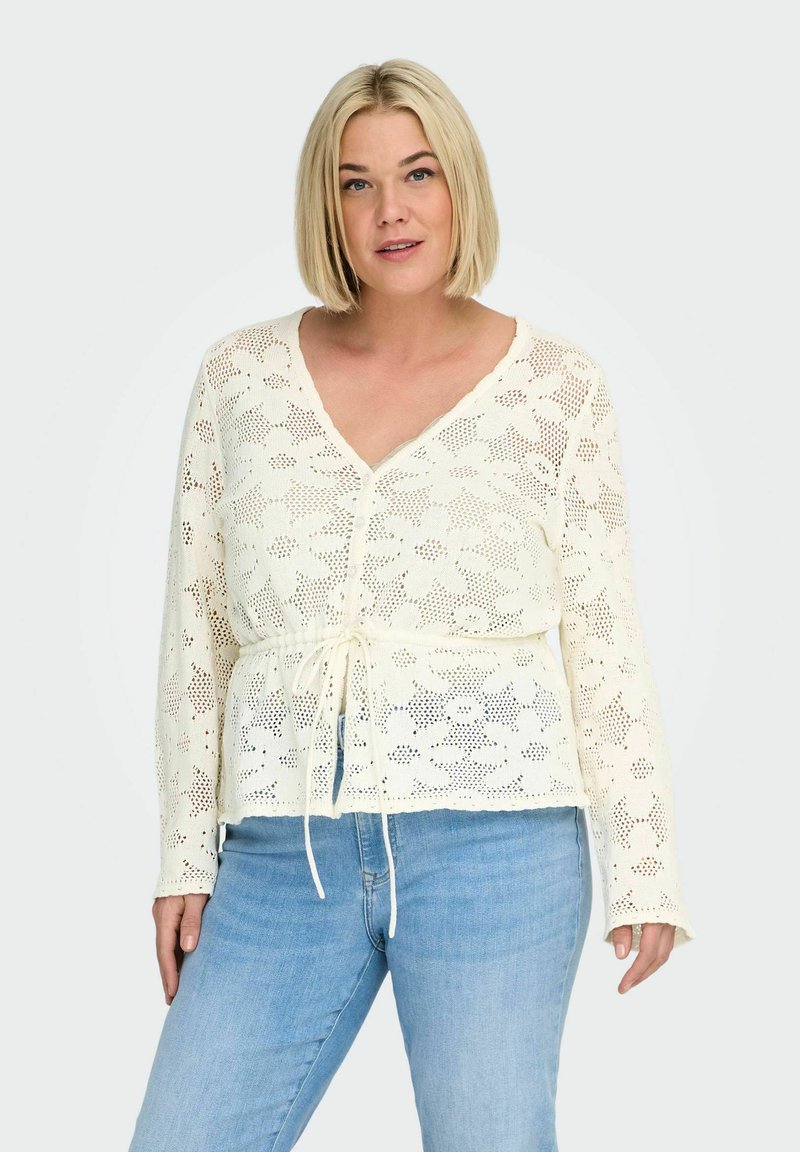 Femme portant un cardigan en dentelle florale couleur crème avec des boutons et un cordon, assorti à un jean bleu clair, debout devant un fond uni.