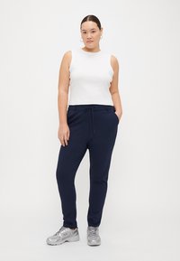 Weißes ärmelloses Oberteil, kombiniert mit marineblauen Jogginghosen mit Kordelzugbund. Modell trägt silberne Sportschuhe und steht vor einem hellen Hintergrund.